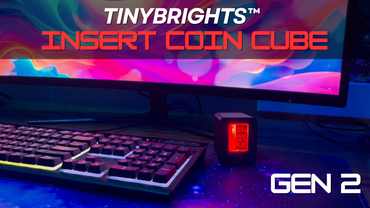 A black textured coin-operated coin slot piggy bank with a red illuminated front panel displaying '25¢ INSERT COIN TO PLAY' in red text sitting on a desk next to a gaming monitor and keyboard.