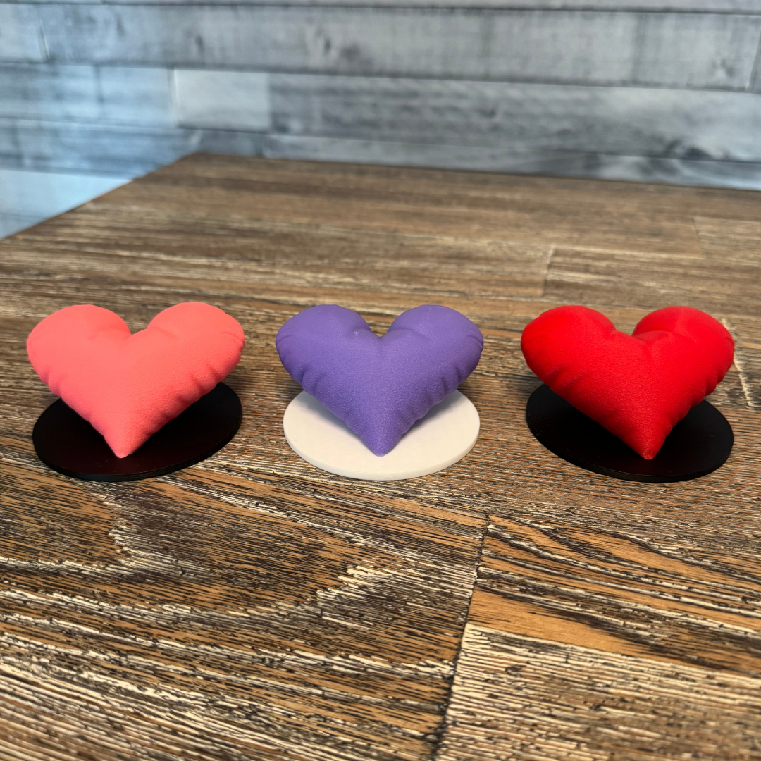 Three 3d printed heart sculptures sitting on a wooden table. Pink, purple, and red heart color options and white and black base color options.