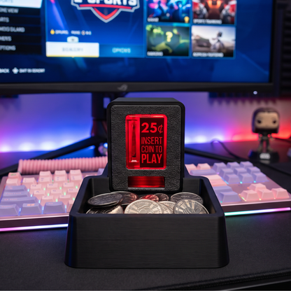 A black coin tray holding several coins, with an Insert Coin Cube displaying '25¢ INSERT COIN TO PLAY' in the center. The setup sits on a desk with a keyboard, monitor, and ambient RGB lighting in the background.
