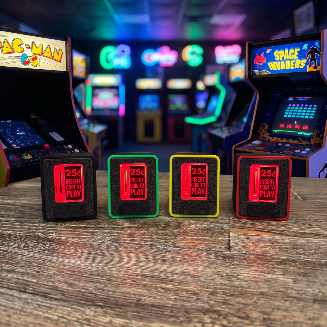 Four retro arcade coin-operated piggy banks lined up on a wooden table in an arcade, with Pac-Man and Space Invaders cabinets blurred in the background.