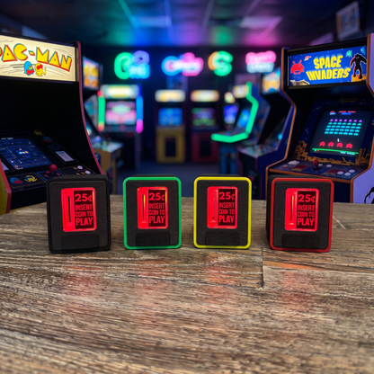 Four retro arcade coin-operated piggy banks lined up on a wooden table in an arcade, with Pac-Man and Space Invaders cabinets blurred in the background.