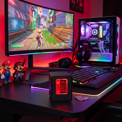 A gaming desk setup in a dimly lit room with a large curved monitor showing a video game, a PC tower with illuminated side panel, RGB keyboard, headphones, and a small black insert coin cube