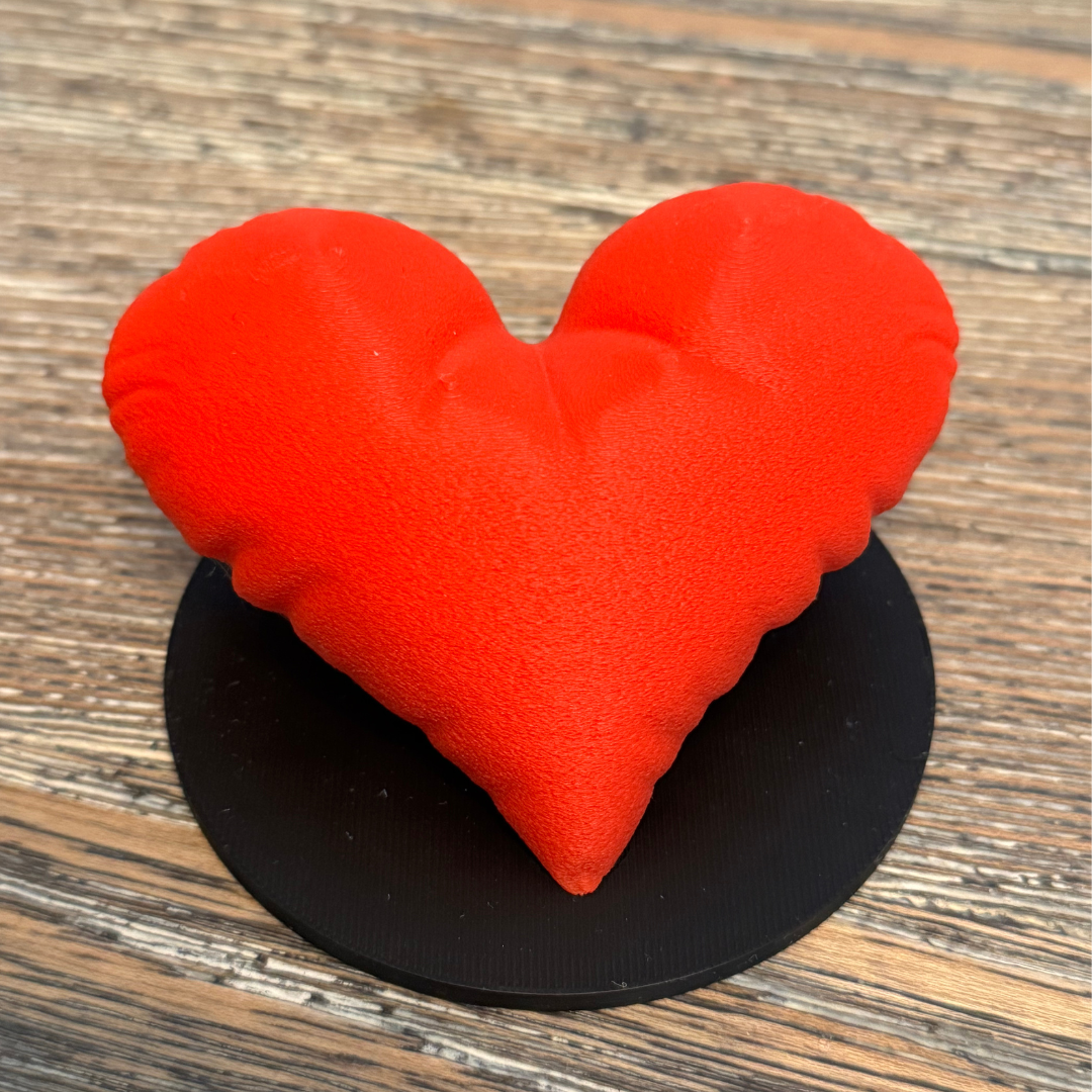 A red 3d printed heart sculpture with a black base sitting on a wooden table.