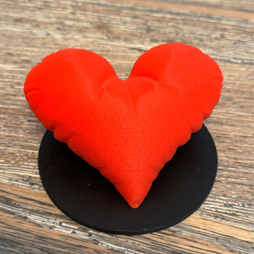 A red 3d printed heart sculpture with a black base sitting on a wooden table.
