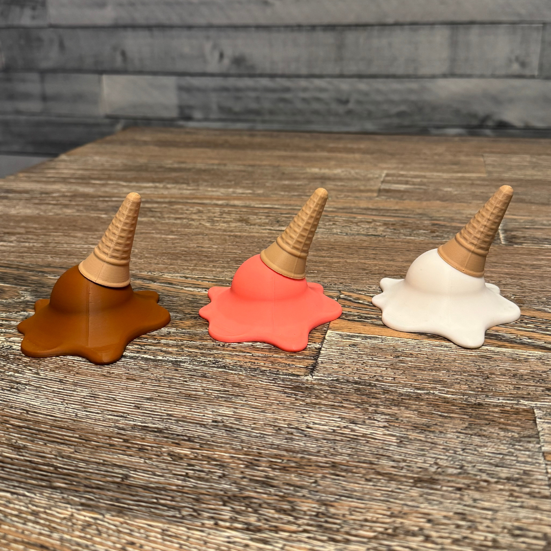 Right side view of three 3d printed melting ice cream cones made by Chronic Concepts sitting on a wooden table. Chocolate, Strawberry, and Vanilla flavored.