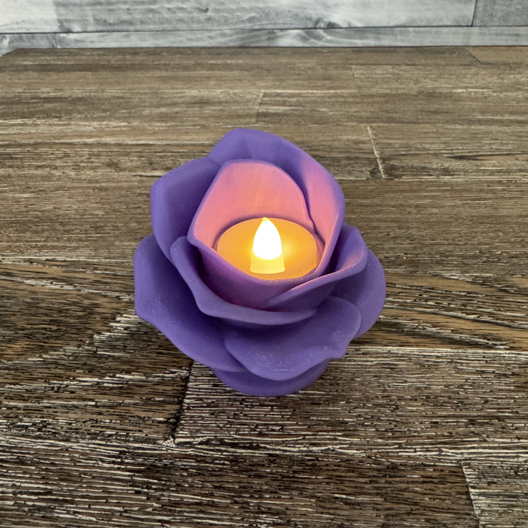 3d printed lilac rose tea light holder with a tea light turned on sitting on a wooden table.