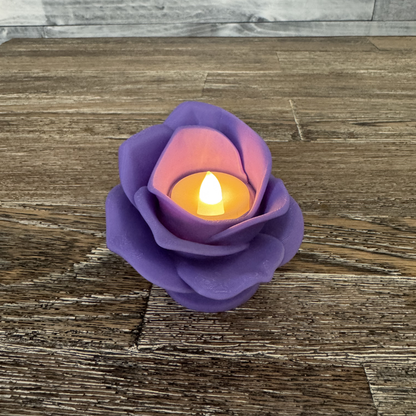 3d printed lilac rose tea light holder with a tea light turned on sitting on a wooden table.