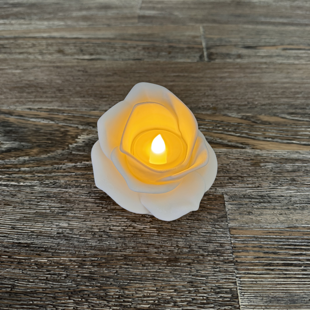 3d printed white rose tea light holder with a tea light turned on sitting on a wooden table.