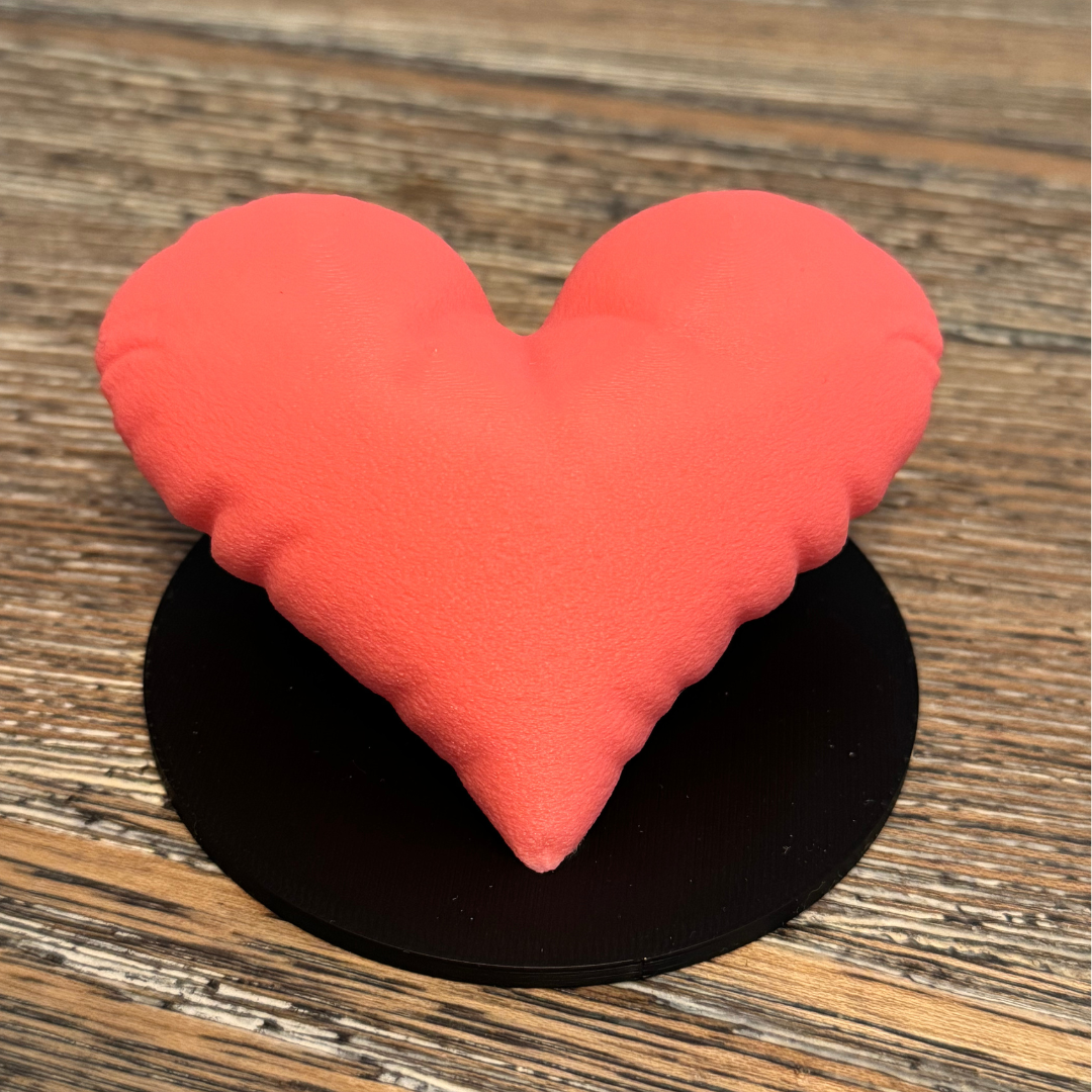 A pink 3d printed heart sculpture with a black base sitting on a wooden table.