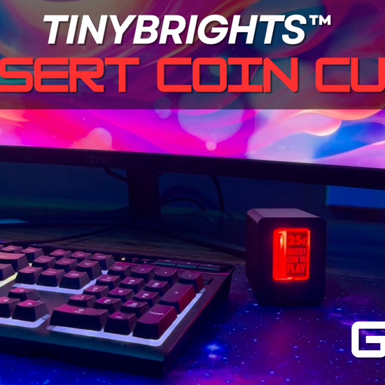 A black textured coin-operated coin slot piggy bank with a red illuminated front panel displaying '25¢ INSERT COIN TO PLAY' in red text sitting on a desk next to a gaming monitor and keyboard.
